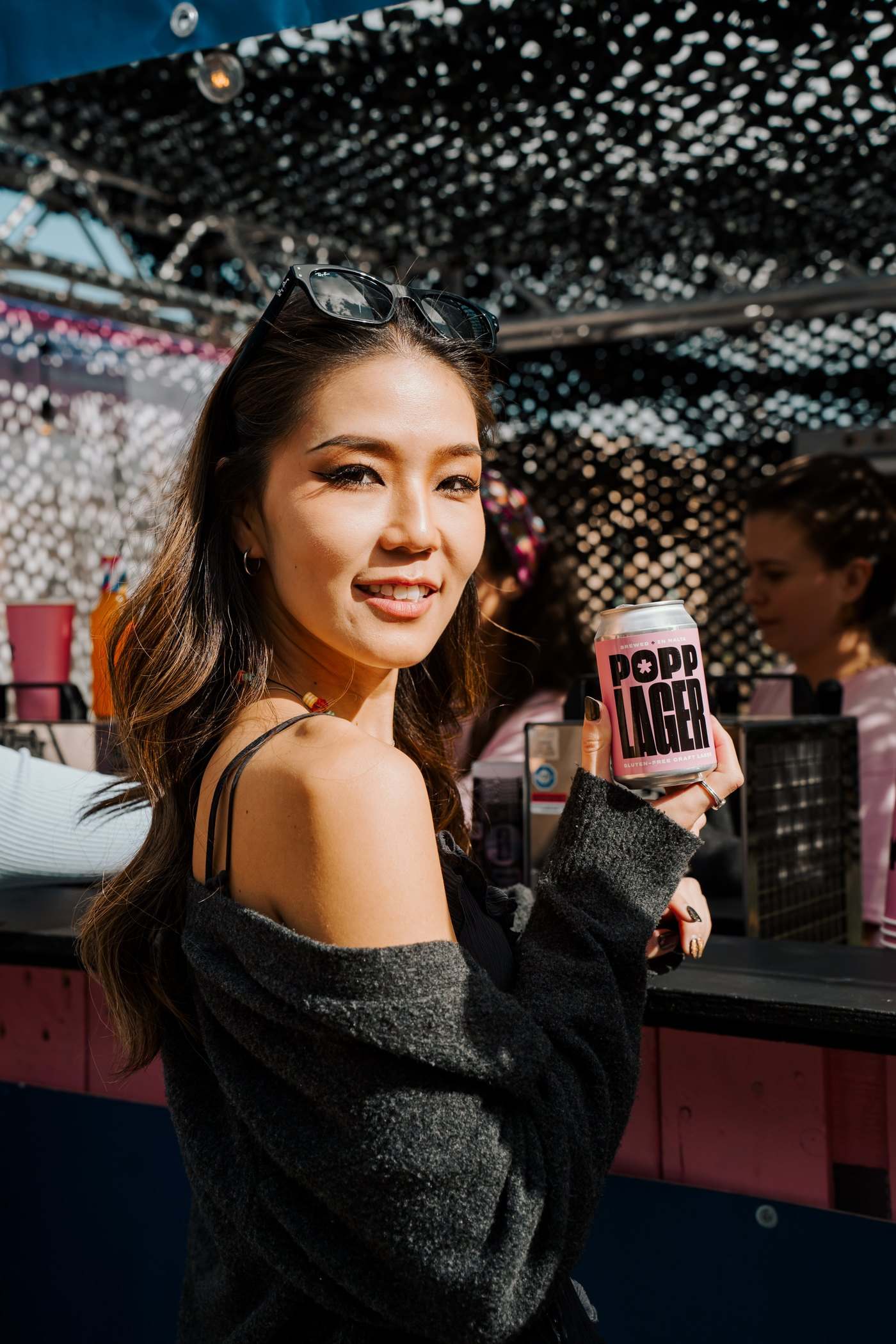 A woman smiling at a bar under a sparkling ceiling, holding a POPP Lager can
