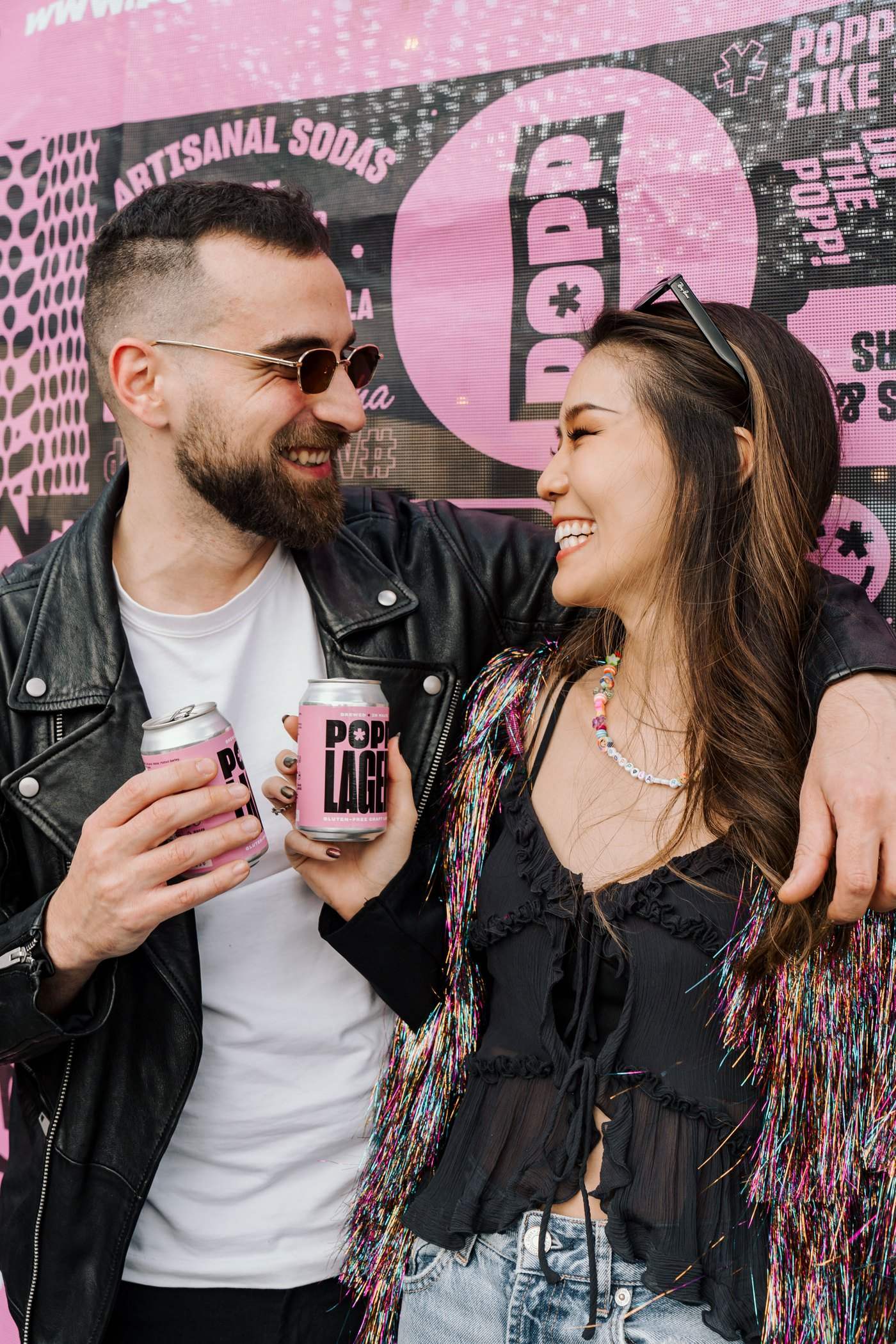 A couple laughing in front of a pink POPP wall, each holding a POPP Lager can