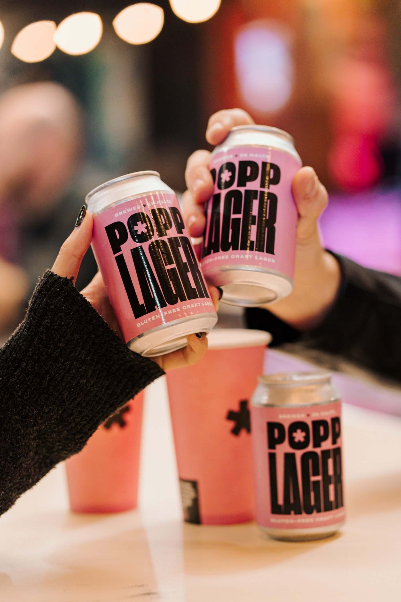 Hands raising pink POPP Lager cans in a cheers over a table with pink cups