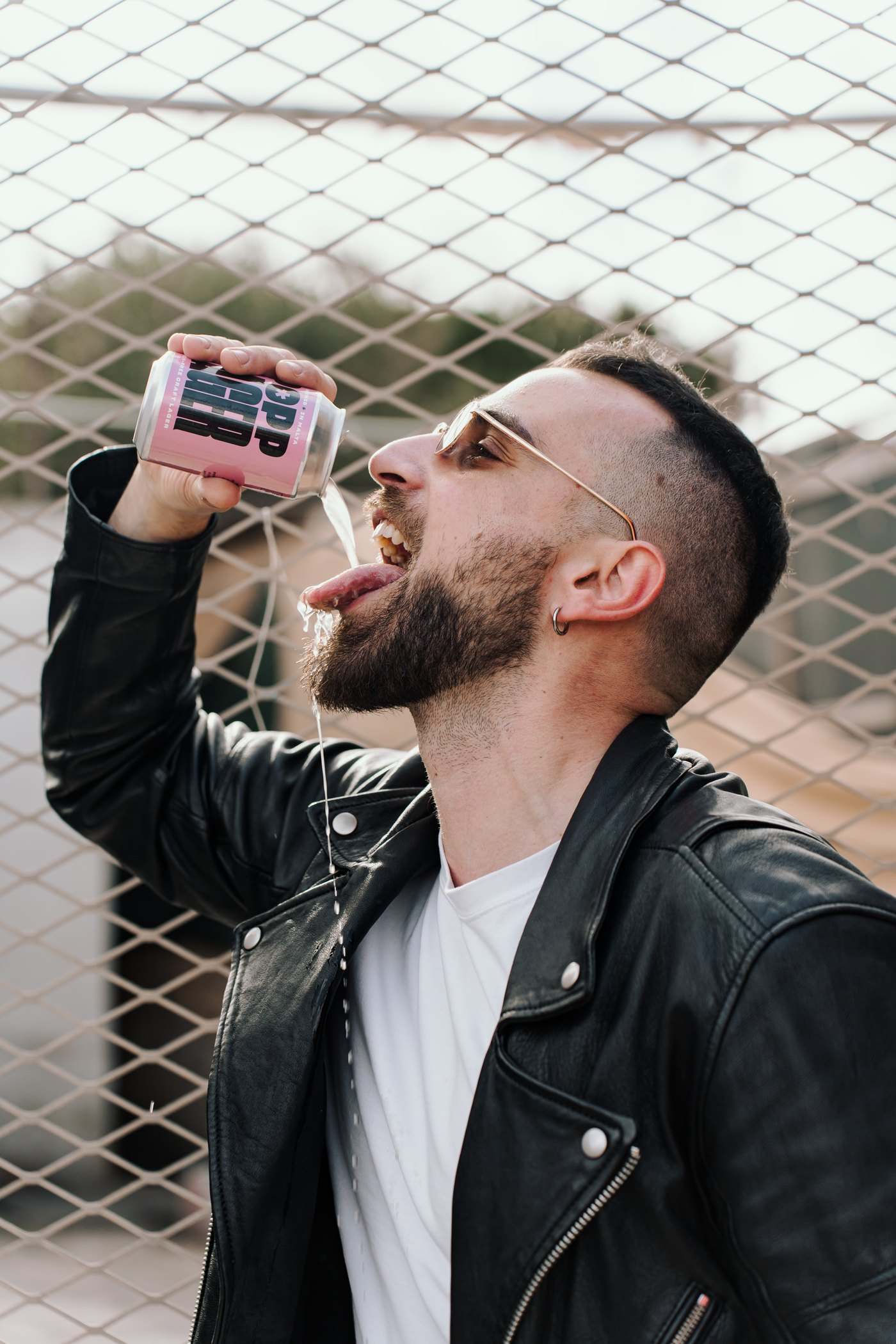 A man in a leather jacket drinking from a POPP Lager can in front of a chain-link fence