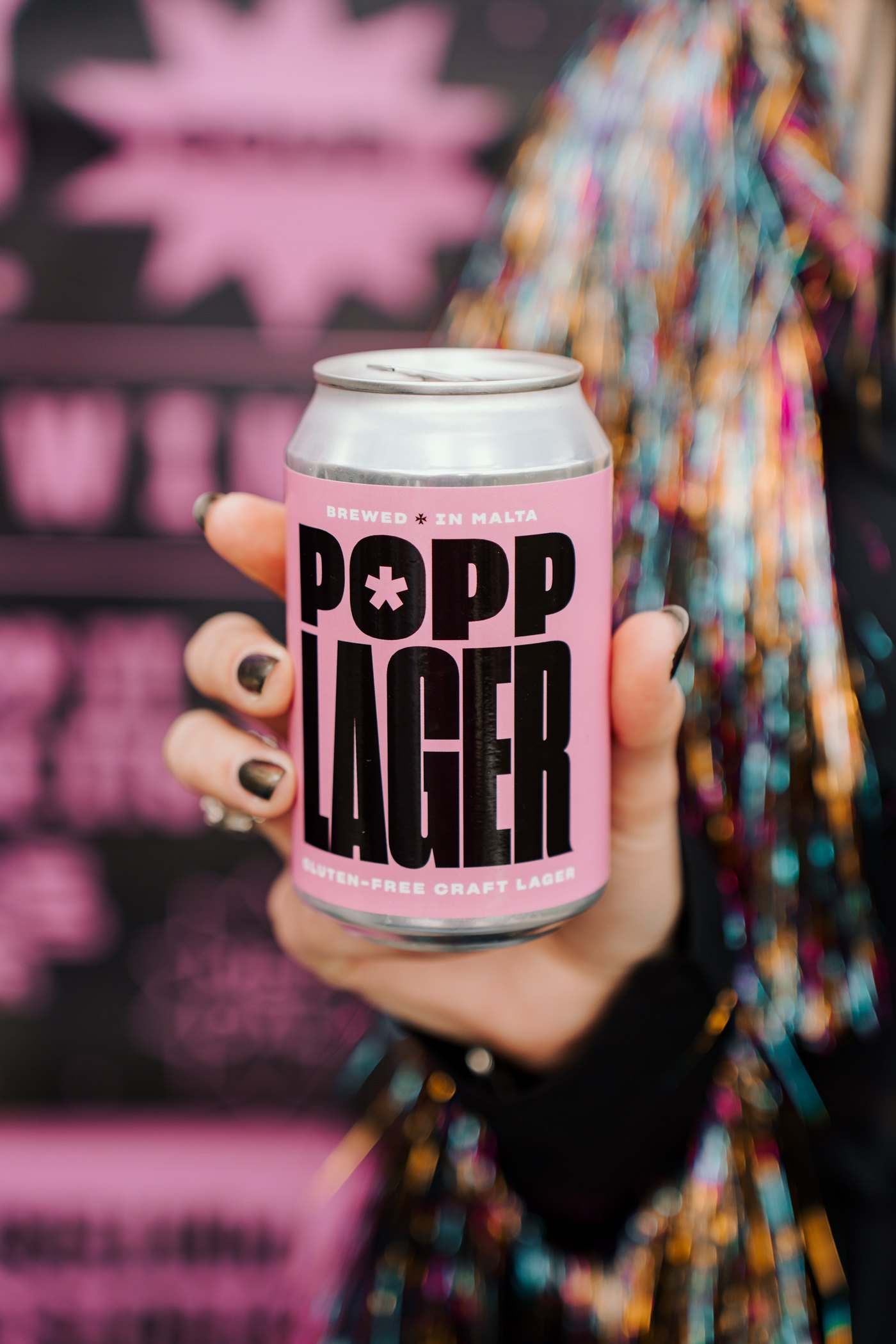 A hand with dark nails holding a POPP Lager can against tinsel and a pink sign