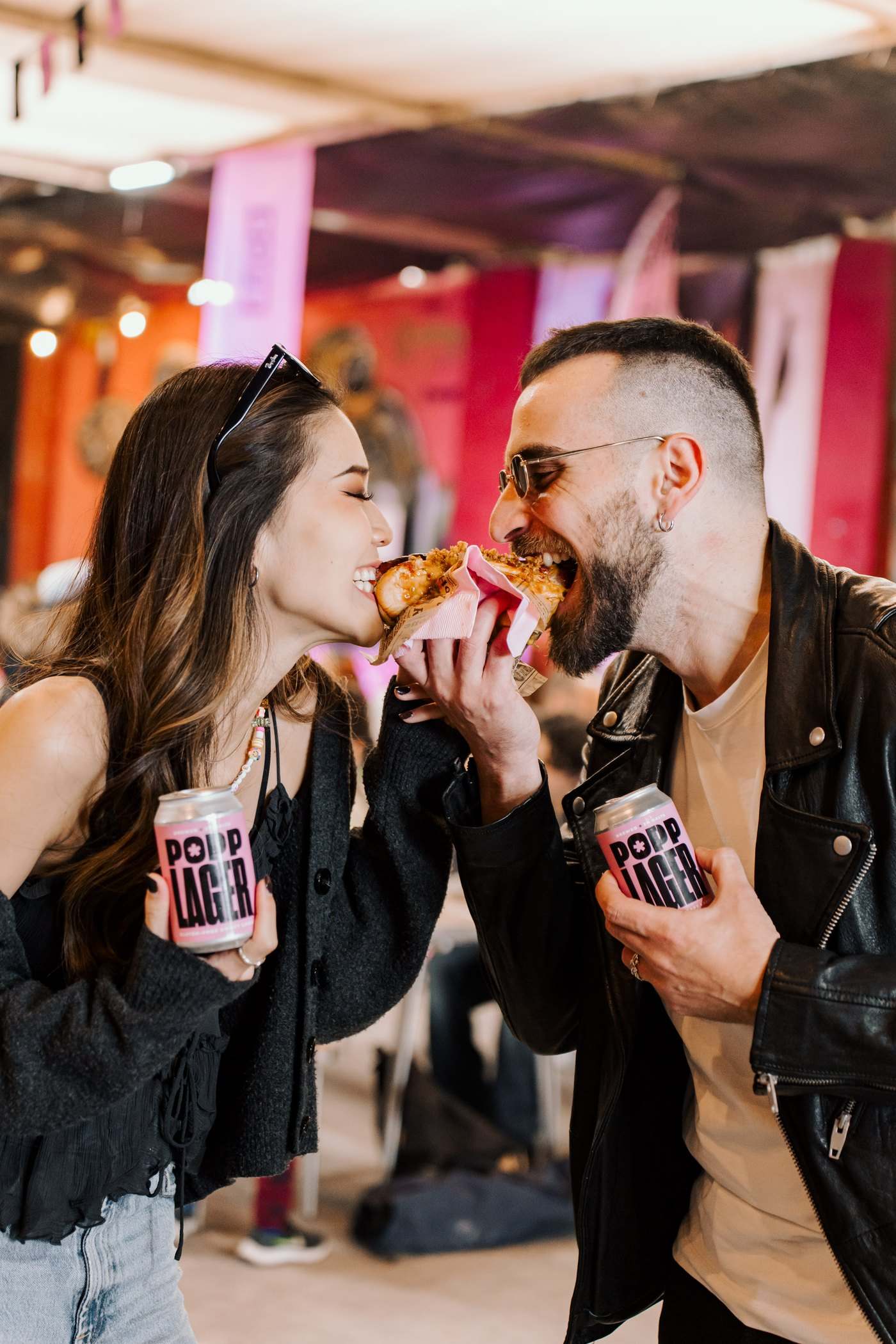 A couple sharing a hot dog at a food hall, both holding POPP Lager cans