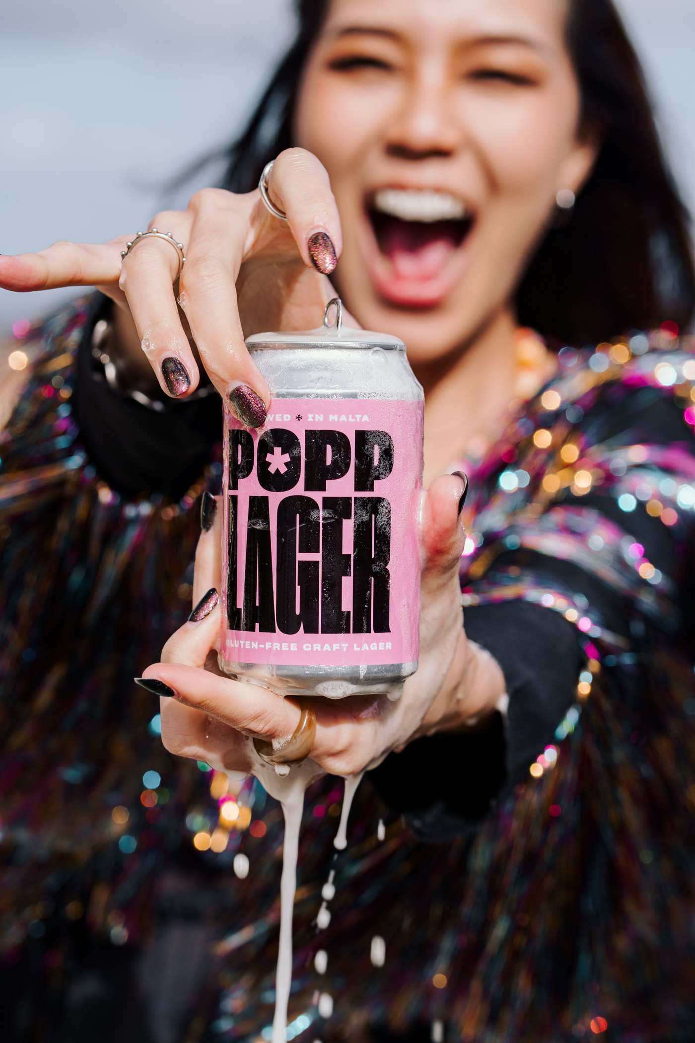 A woman in a sequinned top laughing as she opens a POPP Lager can and it fizzes over
