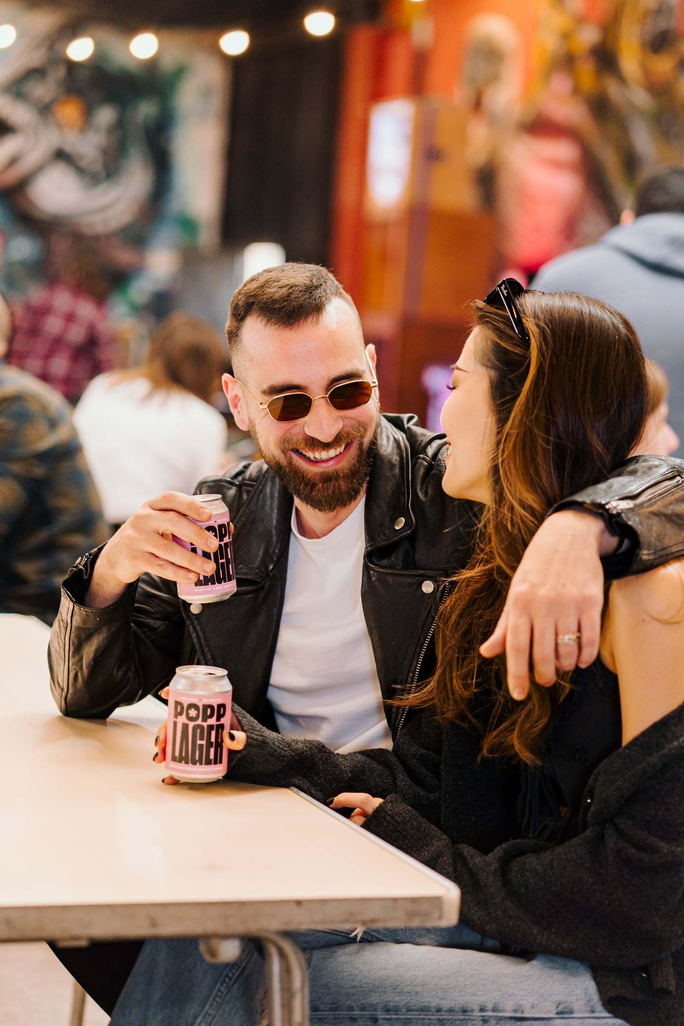 A couple laughing together at a wooden table with POPP Lager cans
